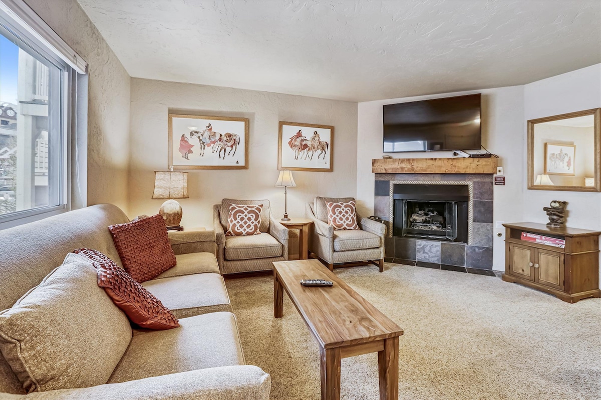 The living room features a stone gas-log fireplace as a focal point, complemented by a large television above a wooden mantel. Comfortable seating includes a plush sofa and two accent chairs, with a wooden coffee table in front. Soft lighting is provided by a lamp, enhancing the cozy ambiance.