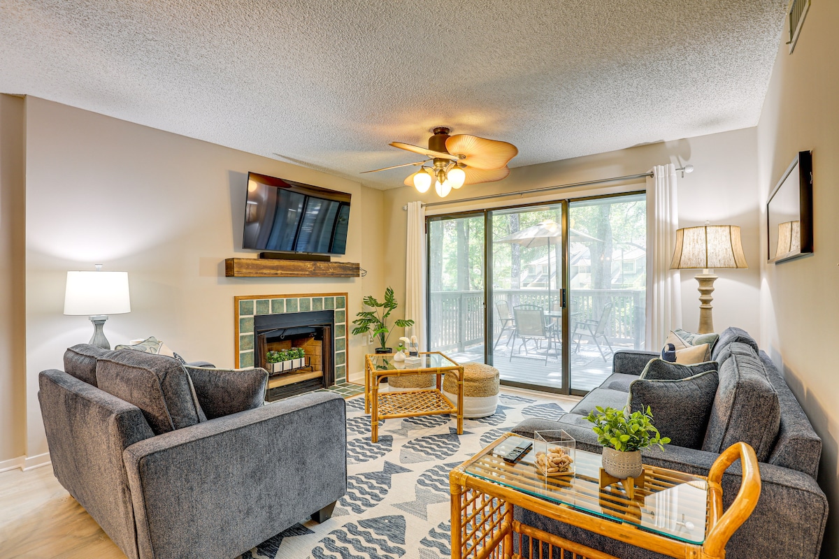 A spacious living area features soft seating arranged around a decorative fireplace. Natural light floods in through large sliding doors that lead to an outdoor deck. A ceiling fan provides air circulation, while decorative elements like plants and a coffee table enhance the inviting layout.