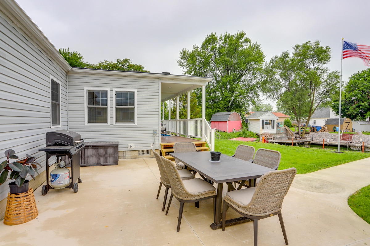 An outdoor patio area is showcased, featuring a dining table surrounded by wicker chairs. A BBQ grill is positioned nearby, and a small plant enhances the space. The well-maintained yard is visible, with a flagpole flying an American flag in the background.