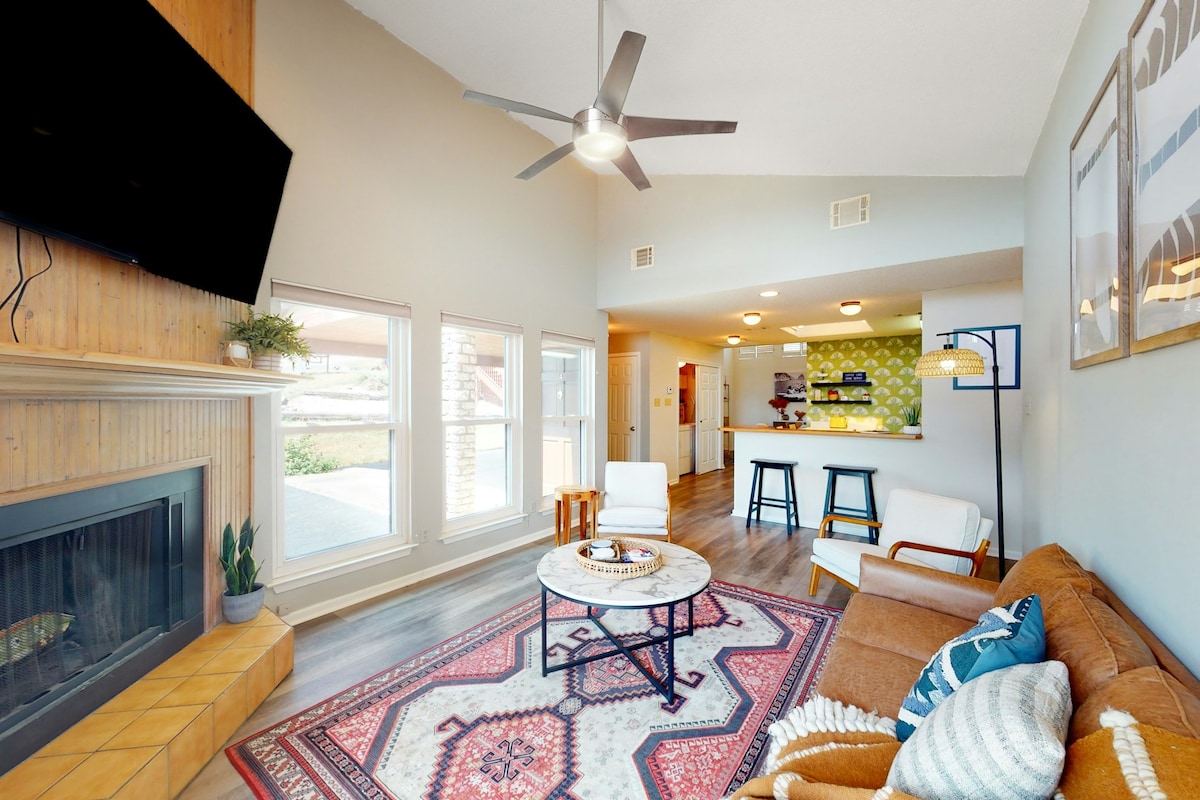A living area features a comfortable sofa and a patterned area rug, complemented by a ceiling fan and a mounted television. Natural light fills the space through large windows, while a well-equipped kitchen is visible in the background.