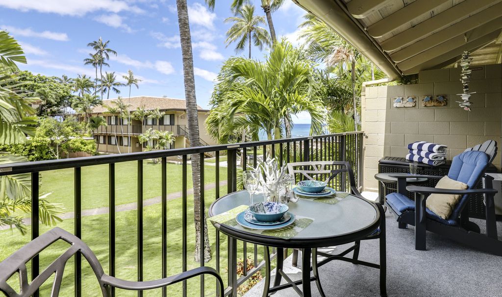 A balcony is showcased with a glass-top table set for two, complemented by blue and white dishware. Adjacent, comfortable chairs provide a space for relaxation. Lush tropical vegetation frames the view, with palm trees and manicured grounds extending toward the ocean in the background.