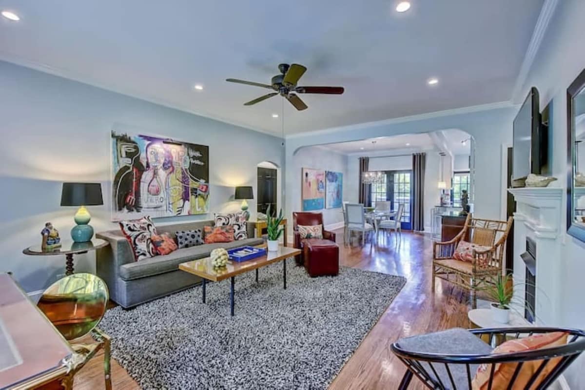 The living room features a spacious layout with a plush area rug and a comfortable sofa adorned with decorative pillows. Original artwork enhances the walls, while natural light flows in from the large windows. A stylish coffee table and various seating options contribute to a welcoming atmosphere.