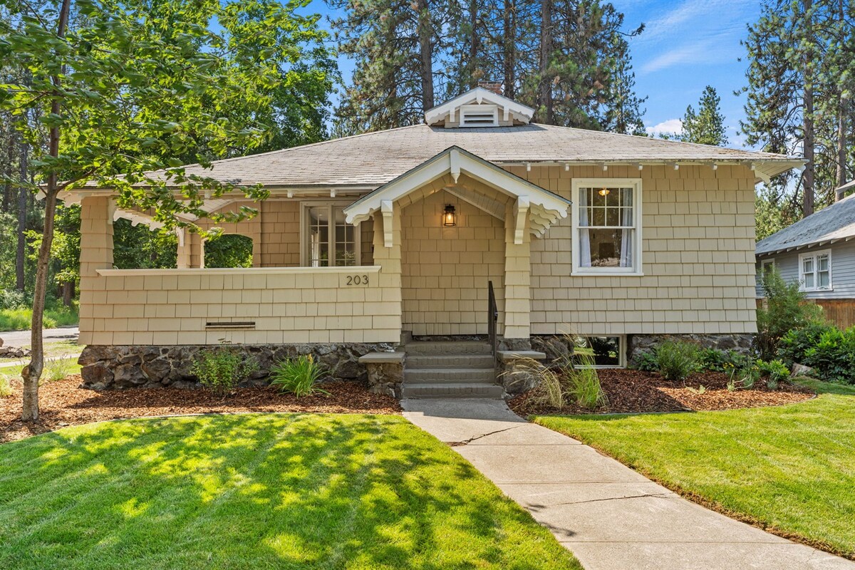 The exterior of the Lorraine House is prominently displayed, featuring a charming sage-colored facade with decorative shingles. A welcoming front porch with steps leads to the entrance, surrounded by lush greenery and well-maintained landscaping. Sunlight gently highlights the grassy area and pathways.