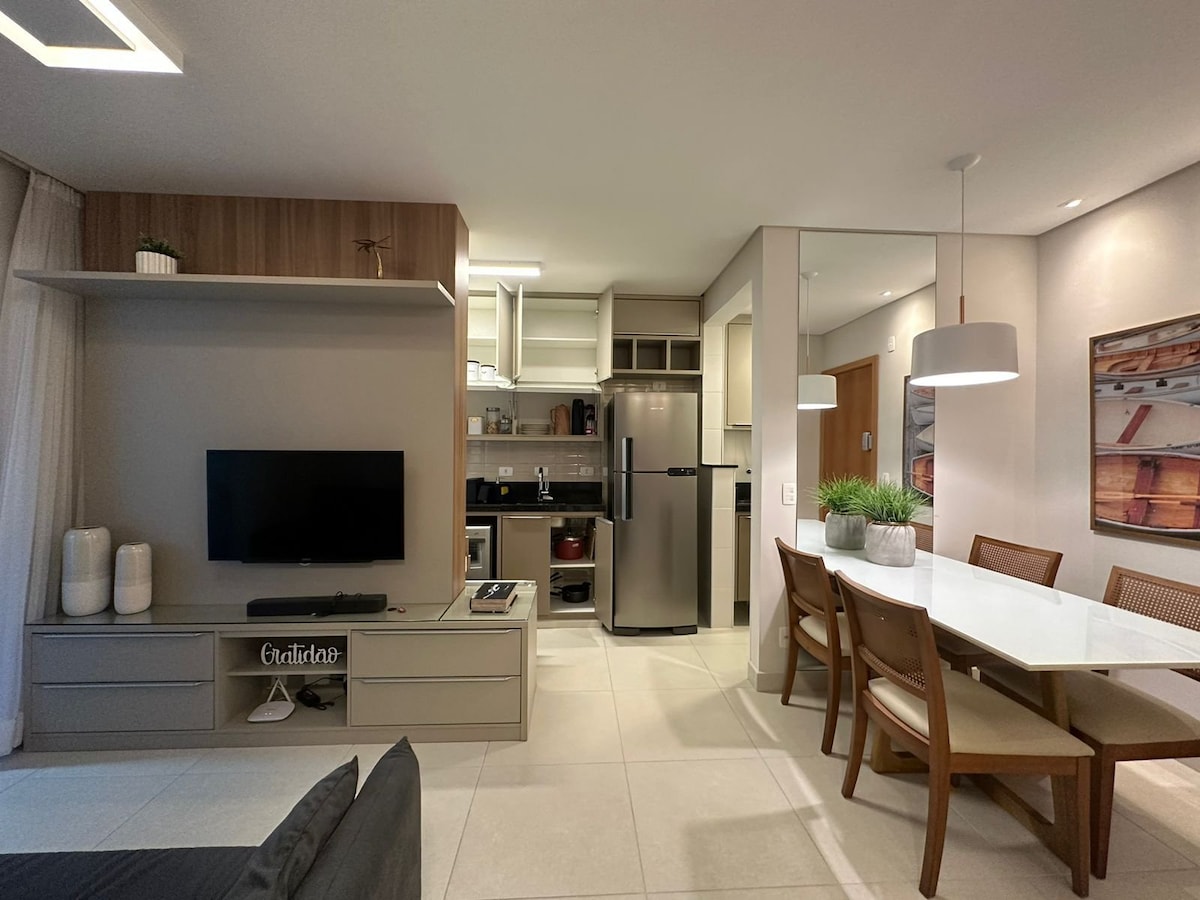 A modern living area features an integrated kitchen with stainless steel appliances and a dining table for six. Light-colored cabinetry and a large flat-screen TV are present. Natural light enters through large windows, complemented by minimalist decor and a touch of greenery.