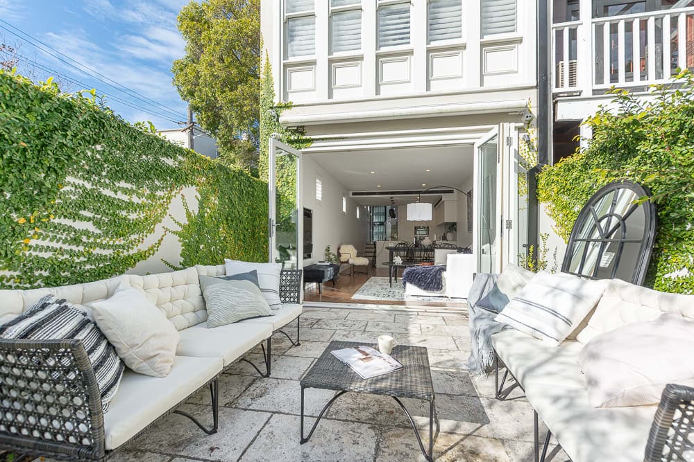 An outdoor seating area is presented with stylish white sofas arranged around a stone patio. Lush green foliage frames the space, providing a serene backdrop. Glass doors open to the interior, creating a seamless transition between indoor and outdoor living.