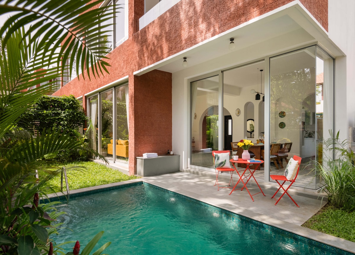 An outdoor patio area is visible, featuring two red chairs and a small table, complemented by lush greenery. A private pool is observed nearby, reflecting sunlight. Large glass doors open into the villa, connecting indoor and outdoor spaces and enhancing the serene ambiance.