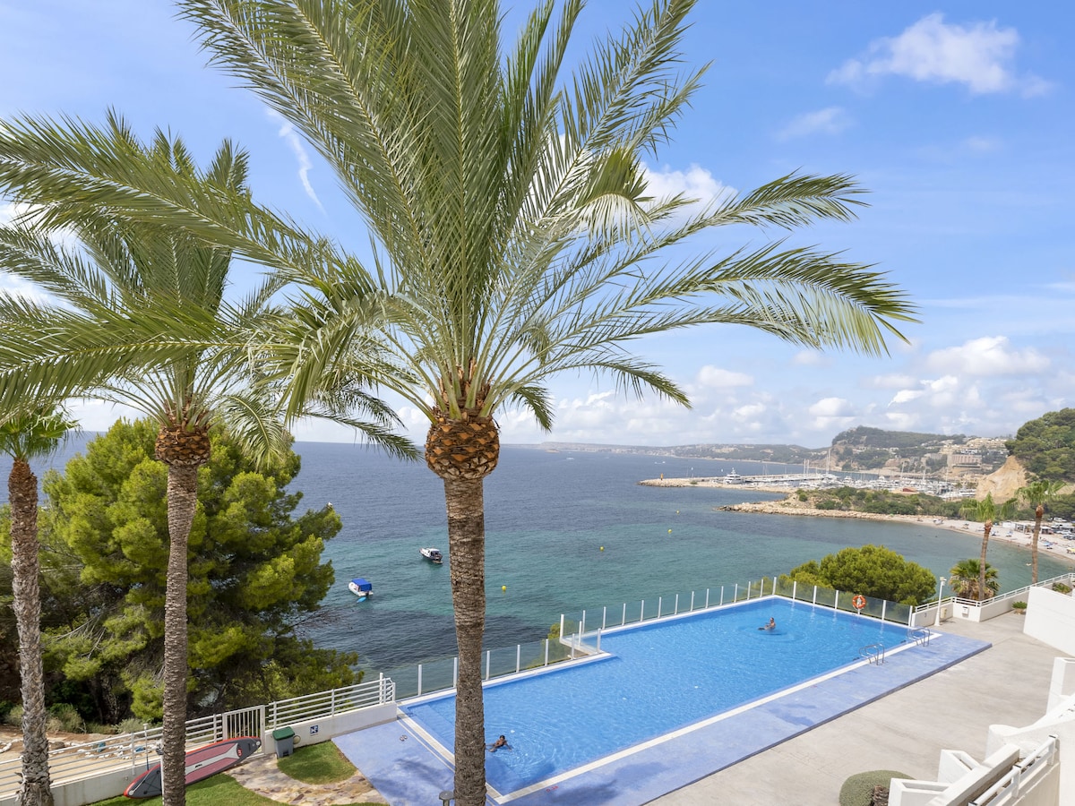 The image captures a swimming pool surrounded by palm trees, with a view of the sea and distant coastline. Gentle waves lap at the water's edge, while boats are anchored nearby. The atmosphere reflects a serene coastal setting, inviting relaxation and enjoyment of the sunny outdoors.