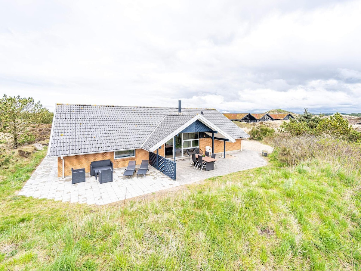 A spacious cottage exterior is surrounded by lush greenery, featuring a partially covered terrace with seating. The large windows provide views of the peaceful landscape, while the characteristic roof adds charm. The area is well-kept, illustrating a tranquil environment sheltered by hilly terrain.