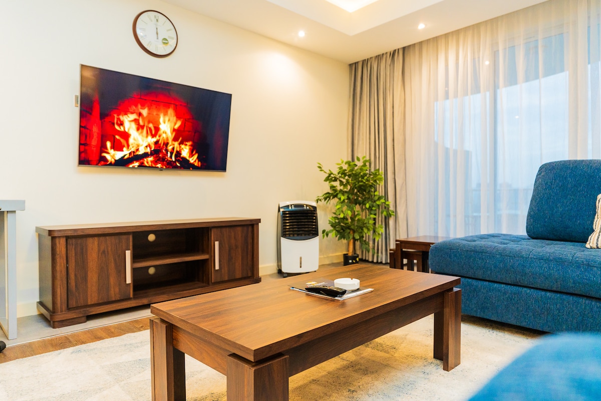 A modern living area is presented with a comfortable blue sofa facing a wooden coffee table. A TV mounted on the wall displays a fireplace scene. A small air conditioning unit and a potted plant add to the ambiance. Natural light filters through sheer curtains.