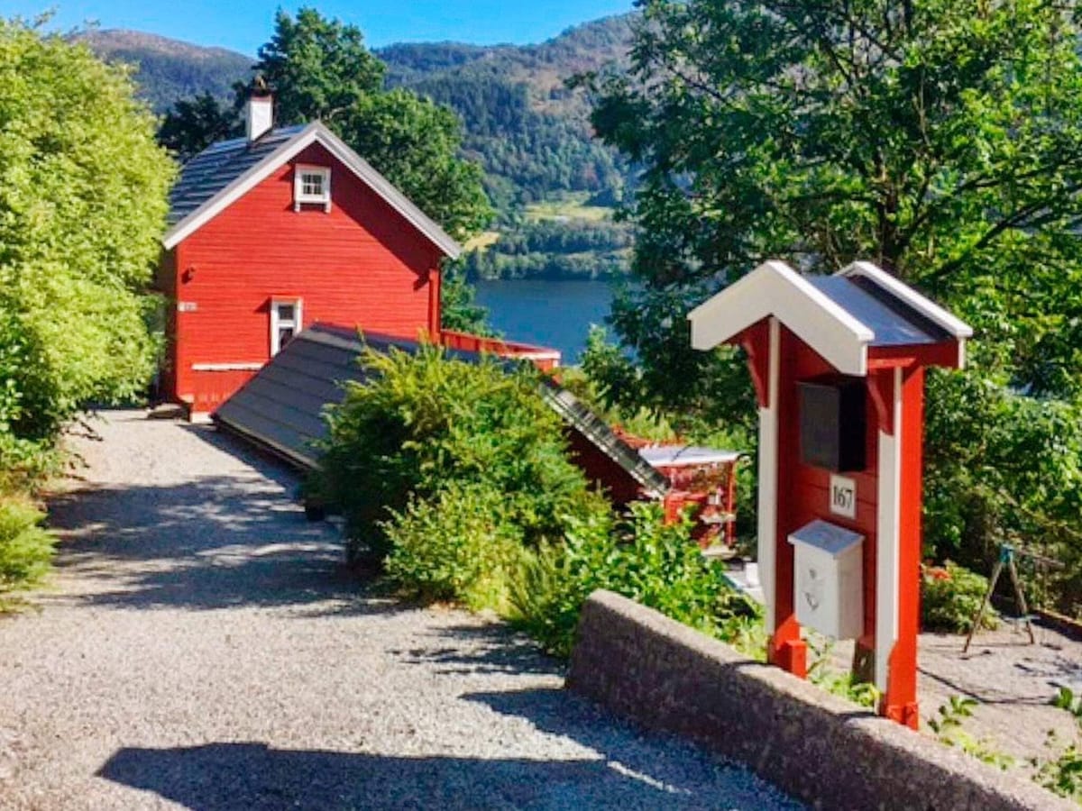 A charming red holiday home is nestled among greenery, with a gravel pathway leading to it. The scenic backdrop features a tranquil body of water and hills, enhancing the peaceful surroundings. A mailbox stands at the edge of the property, contributing to the inviting atmosphere.