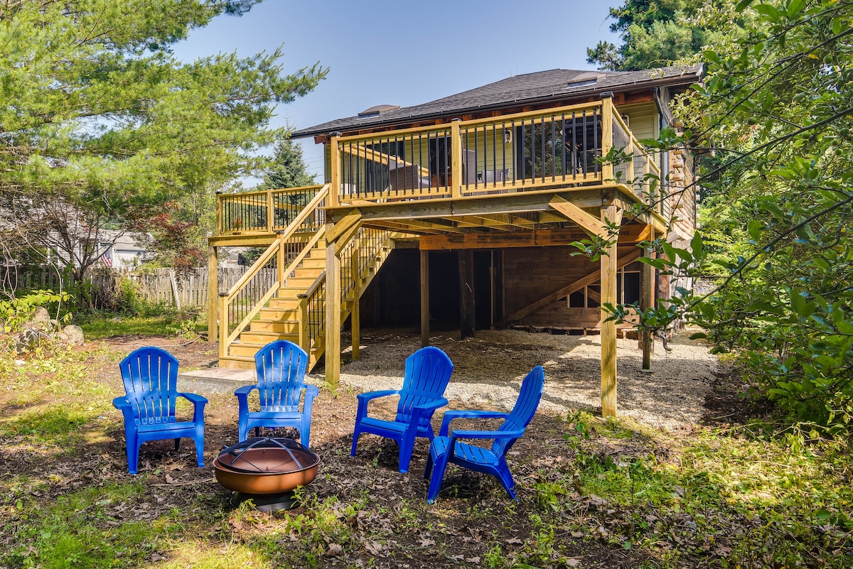 An outdoor area features a fire pit surrounded by four blue Adirondack chairs. A wooden deck is elevated above, providing access via a staircase. Natural greenery and gravel paths enhance the serene setting.