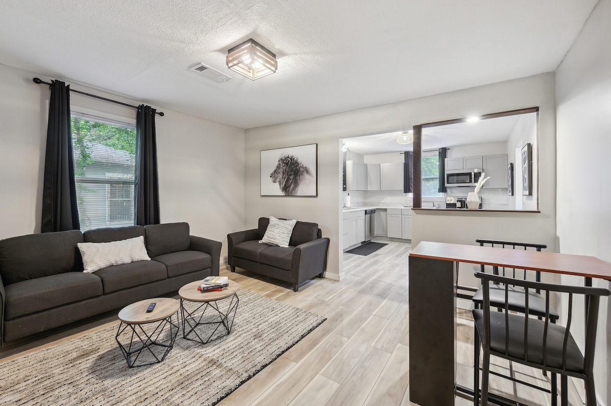 A living area features two dark sofas positioned across from each other, enhancing comfort and relaxation. A coffee table with a geometric design stands on a textured area rug. A dining counter is visible in the background, leading into a well-lit kitchen space.