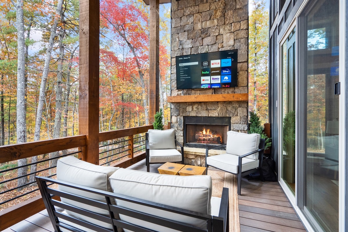A covered porch features a stone fireplace, surrounded by comfortable seating for relaxation. A large television is mounted above the fireplace, offering entertainment options. The scenic backdrop displays vibrant fall foliage through trees, creating a picturesque outdoor space for unwinding.