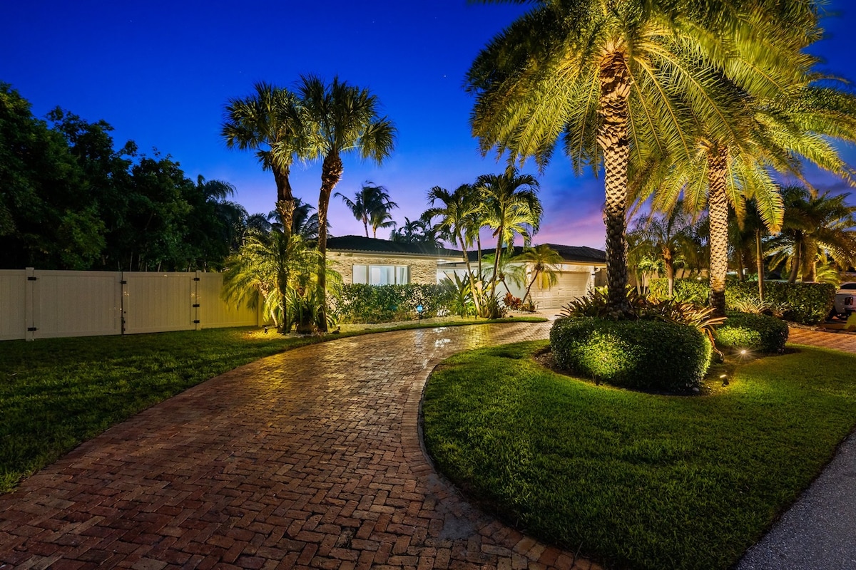 The exterior of the property features a landscaped pathway illuminated by soft lighting, bordered by tall palm trees and lush greenery. A well-maintained lawn enhances the inviting curb appeal, with the residence appearing under a dusk sky.