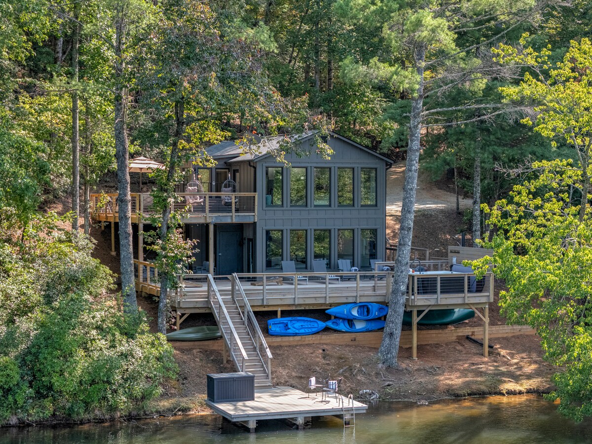 A modern two-story house is situated along the edge of a tranquil lake, surrounded by lush greenery. Large windows face the water, allowing natural light to flood the interior. A wooden deck extends from the house, providing access to the private dock and kayaks.