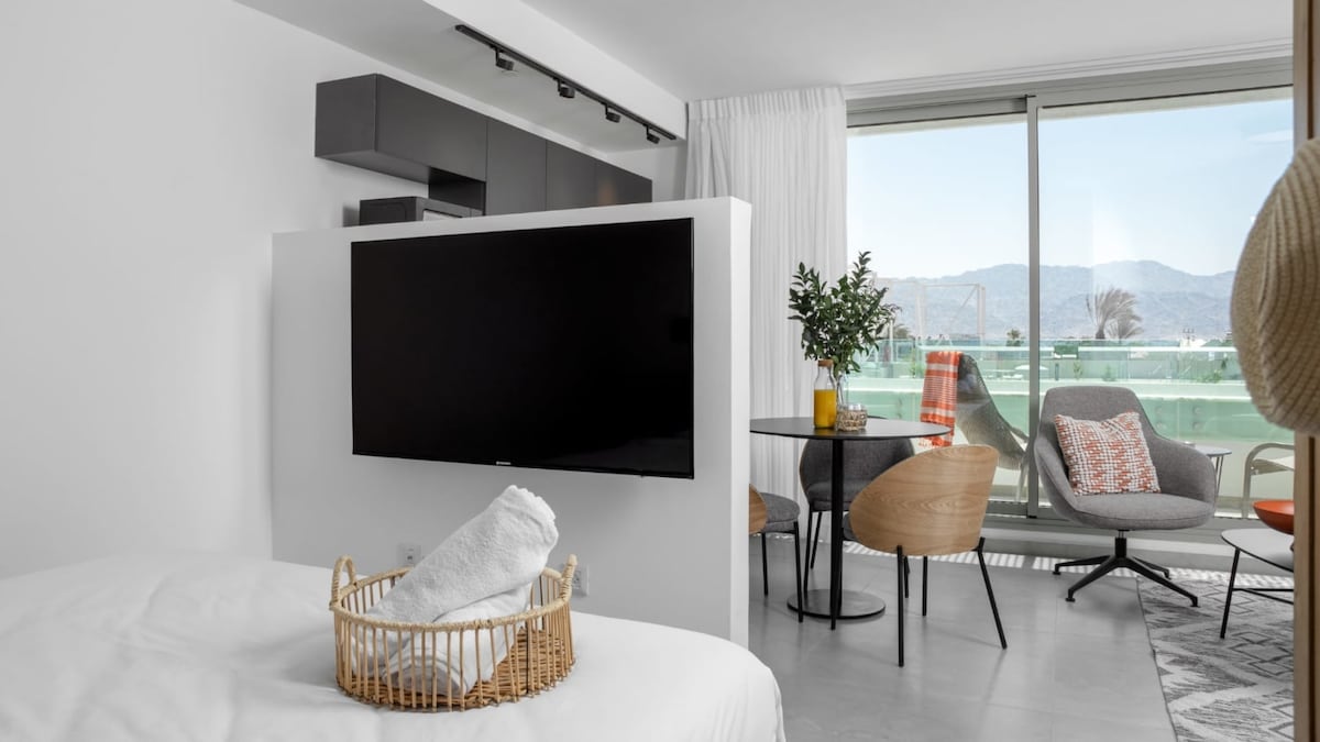 A bright and airy studio space features a modern design with a large window overlooking a serene view. A comfortable seating area includes a round table and stylish chairs, while a wall-mounted television is positioned for easy viewing. A towel is neatly arranged in a woven basket.