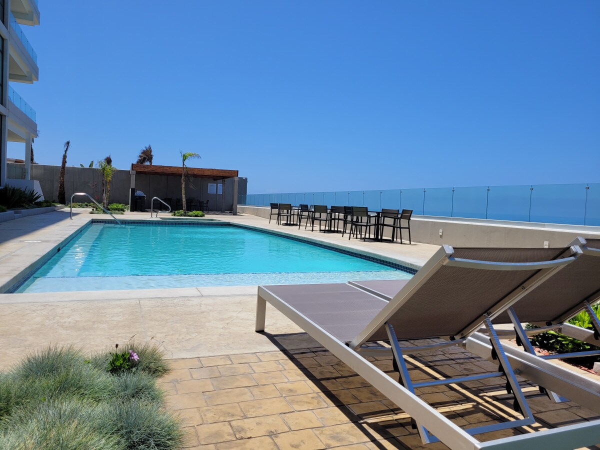 A shared pool area is observed, featuring a clear blue swimming pool surrounded by a spacious deck. Lounge chairs are positioned on one side, while a shaded BBQ area with seating is visible in the background. The ocean can be seen in the distance under a clear blue sky.