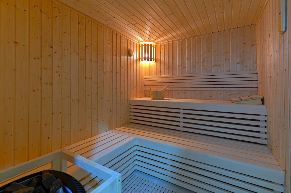 A wooden sauna features neatly arranged wooden benches and walls, complemented by soft lighting from a wall-mounted fixture. A wooden bucket and ladle are present, providing an inviting space for relaxation and warmth.