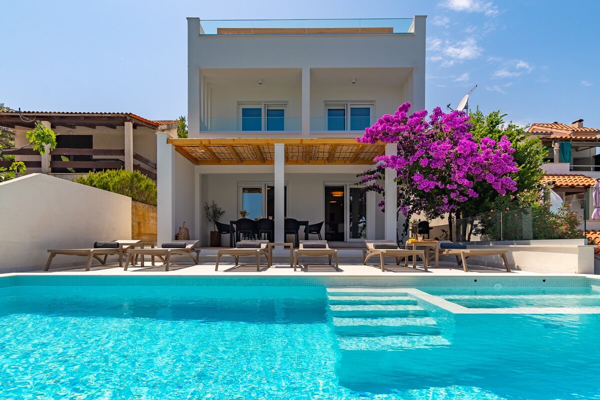 A stylish villa is showcased with a spacious terrace and a private heated pool. Sun loungers are positioned around the pool area, with vibrant purple bougainvillea adding a touch of color. The structure has large glass doors, providing a view of the outdoor seating space.