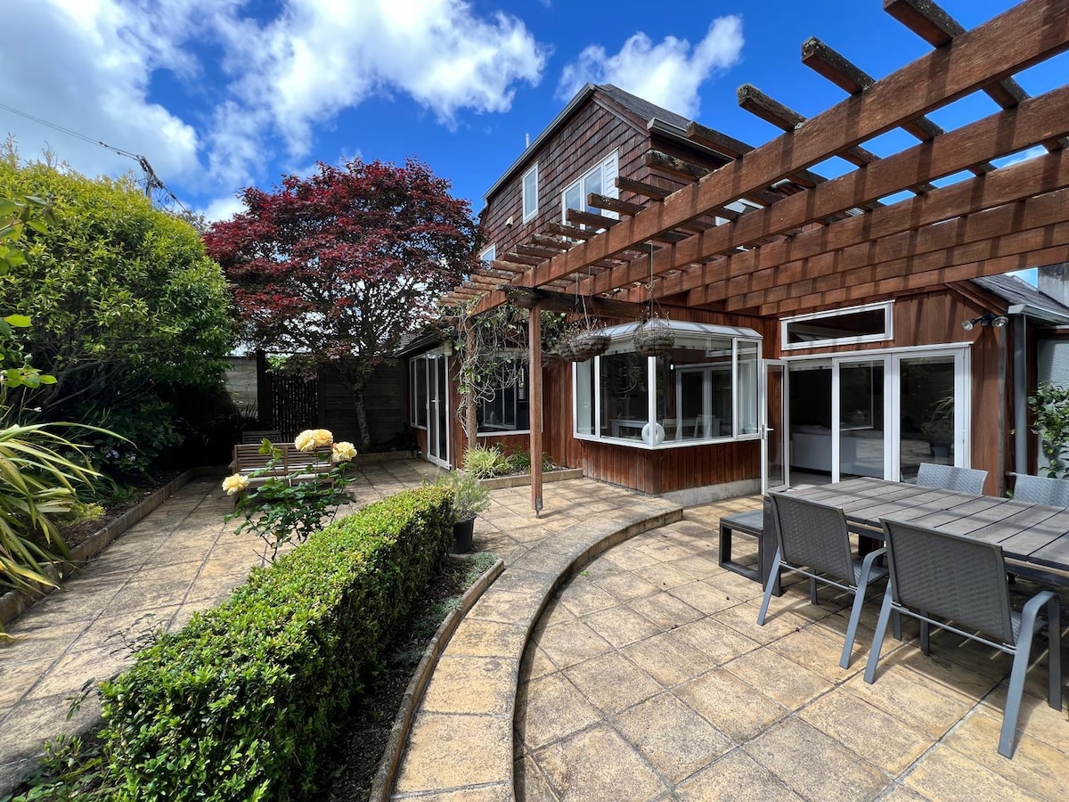 The sun-drenched courtyard features a stone patio surrounded by lush greenery and flowering plants. A spacious outdoor dining table is positioned beneath a wooden pergola, providing shade and a sense of tranquility. Bright blue skies and scattered clouds enhance the inviting outdoor space.