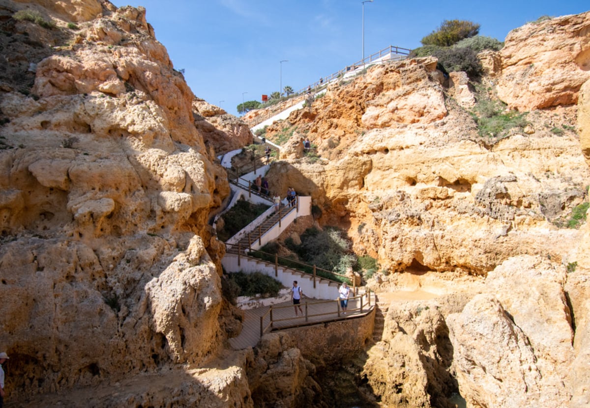 A scenic pathway winds through rocky terrain, leading towards the top of the cliffs. Wooden steps are visible along the path, allowing for easy navigation. People can be seen enjoying the walk, with greenery accentuating the natural landscape.