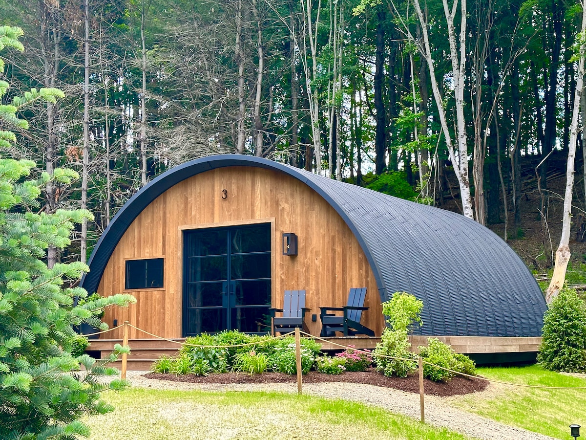 An elegantly designed Wellness Hut showcases a curved shape with a wooden facade. Two dark blue chairs are positioned on the front porch. Lush greenery and a well-maintained path frame the hut, while trees provide a natural backdrop.