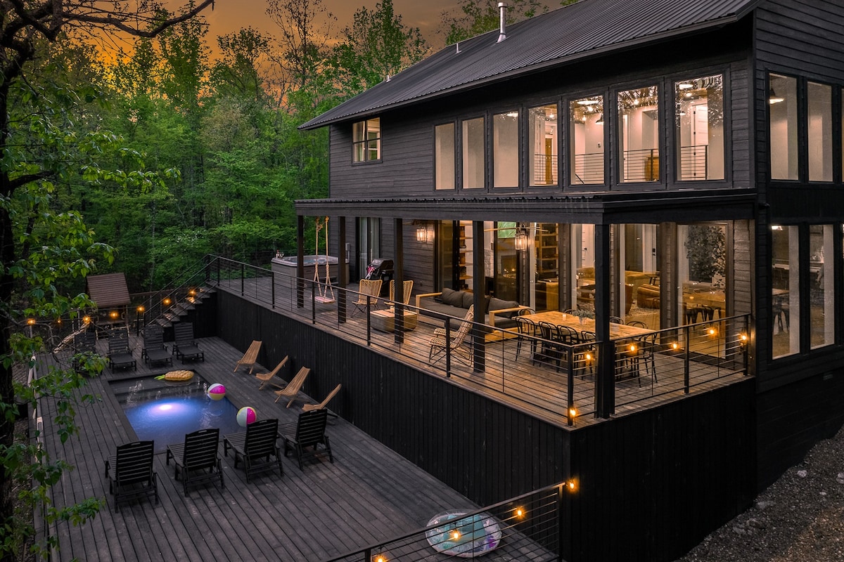 A two-story cabin enveloped by lush greenery features a spacious outdoor deck with lounge chairs and a private pool. Large glass doors provide views into the bright interior, where modern furnishings and a welcoming atmosphere are visible. Soft lighting enhances the evening ambiance.