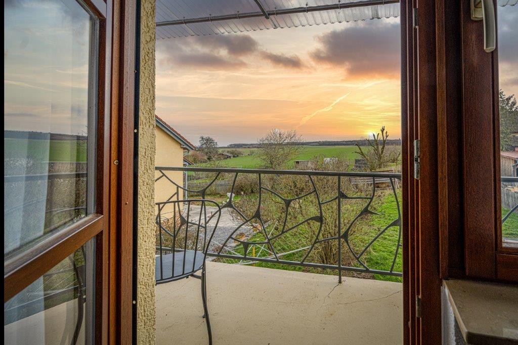 A balcony view is presented, showcasing an expansive landscape with green fields under a sunset sky. A small round table and a chair are positioned on the balcony, framed by an elegant railing. The scene illustrates tranquility and a direct connection to nature.