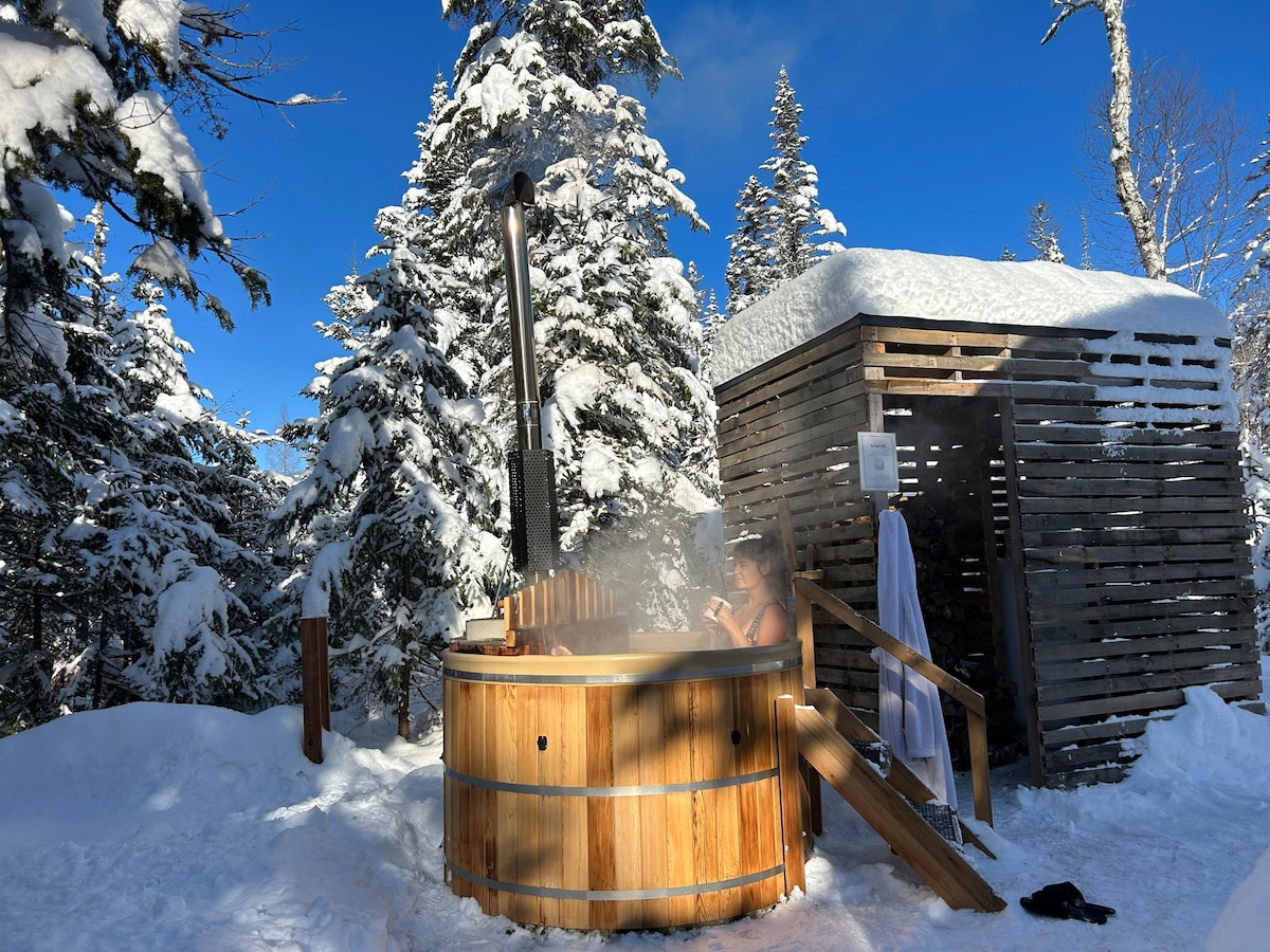 A wood-fired spa is surrounded by pristine snow and towering evergreen trees under a clear blue sky. The spa's wooden exterior complements the natural environment, while steam rises gently, suggesting warmth and relaxation amidst the winter landscape.