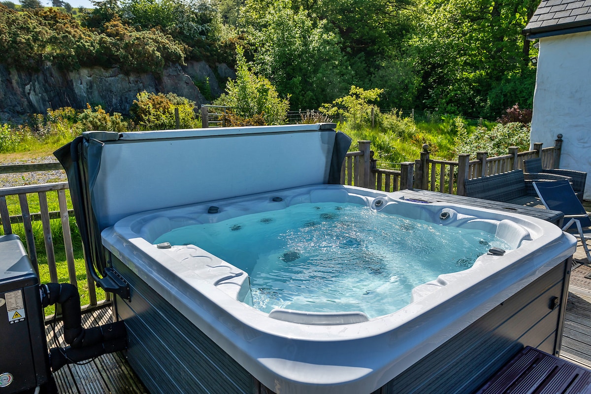 A modern hot tub is situated on a wooden deck, overlooking lush greenery and natural surroundings. Water bubbles gently in the tub, inviting relaxation, while the deck features space for seating.