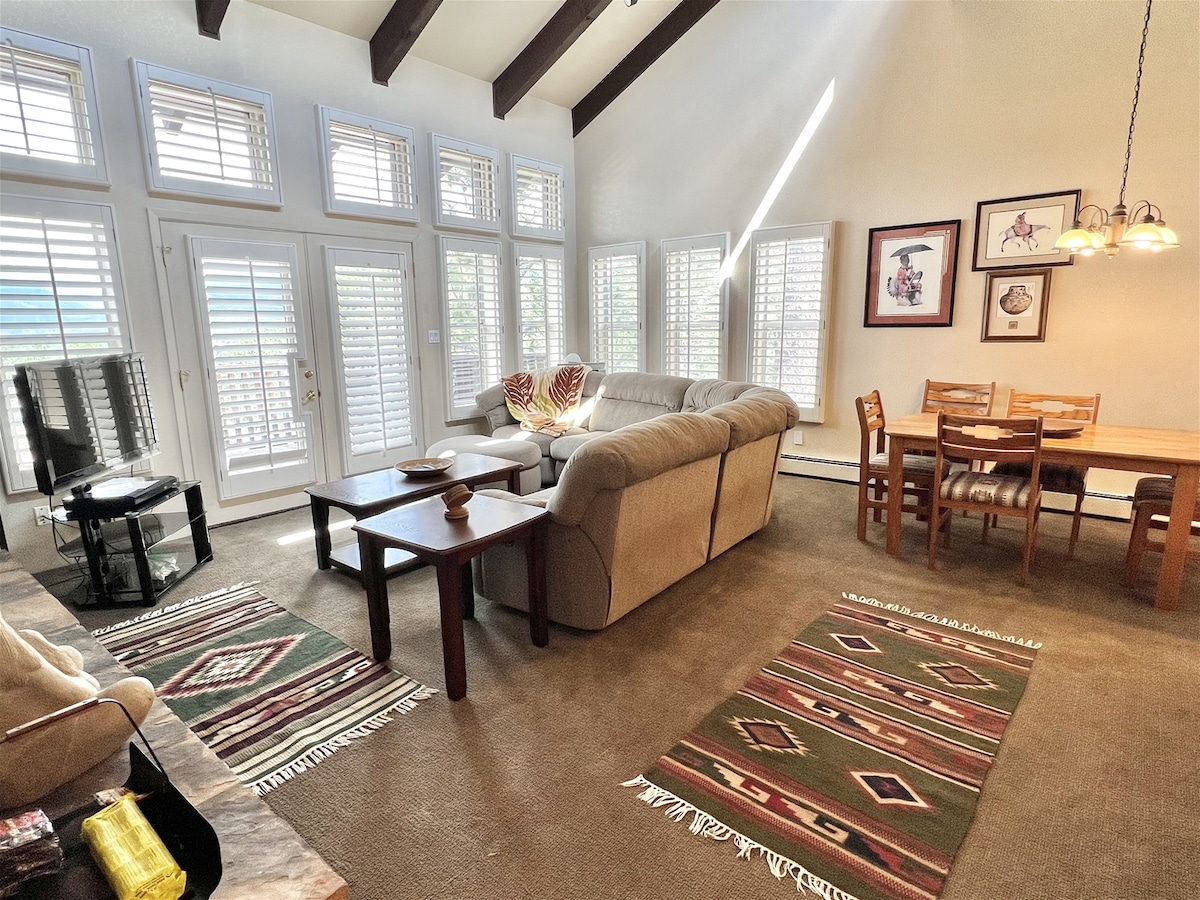 A spacious living and dining area features high ceilings with beams. The room includes a sectional sofa, a coffee table, and a dining table with chairs. Natural light filters through multiple shuttered windows, with decorative area rugs adding warmth to the space.