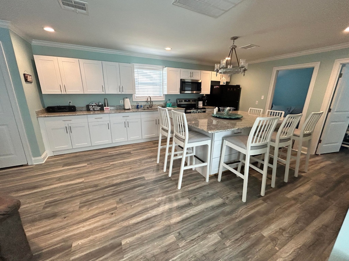 A modern kitchen space is shown, featuring white cabinetry and a large island with seating for six. Stainless steel appliances, including a refrigerator and microwave, are visible. Neutral-colored flooring complements the room, which is illuminated by overhead lighting.