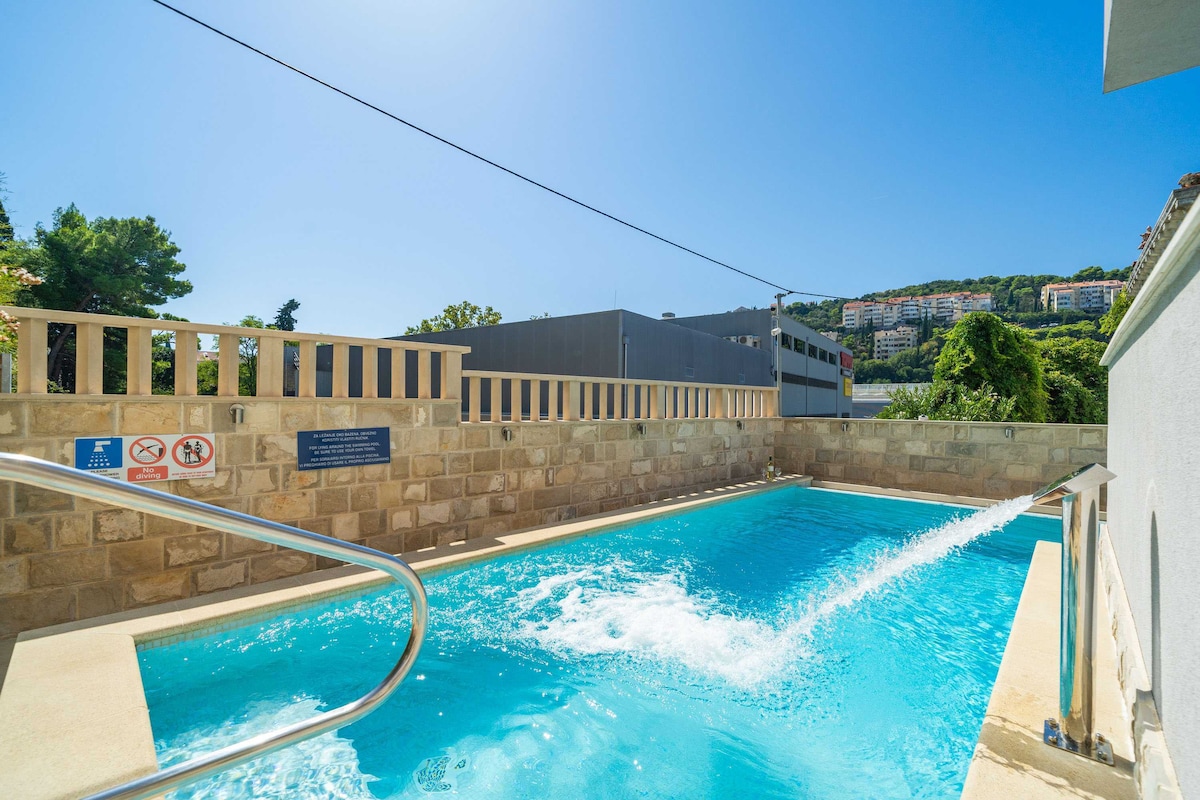 The outdoor seasonal swimming pool is featured, with clear blue water gently disturbed by a fountain. Surrounding stone walls provide privacy, while lush greenery and distant hills create a natural backdrop. Safety signs are displayed, and the pool area is well-maintained.