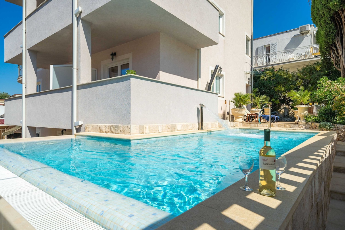 A clear swimming pool is framed by a stone deck, with a bottle of white wine and glasses placed on the edge. The pool reflects the blue sky, while the modern building stands in the background, surrounded by greenery and outdoor seating.