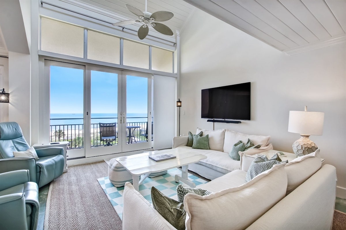 An inviting living area features a large sectional sofa adorned with various cushions, complemented by two recliners. A flat screen TV is mounted on the wall. Sliding glass doors allow natural light and provide a view of the ocean and outdoor space.