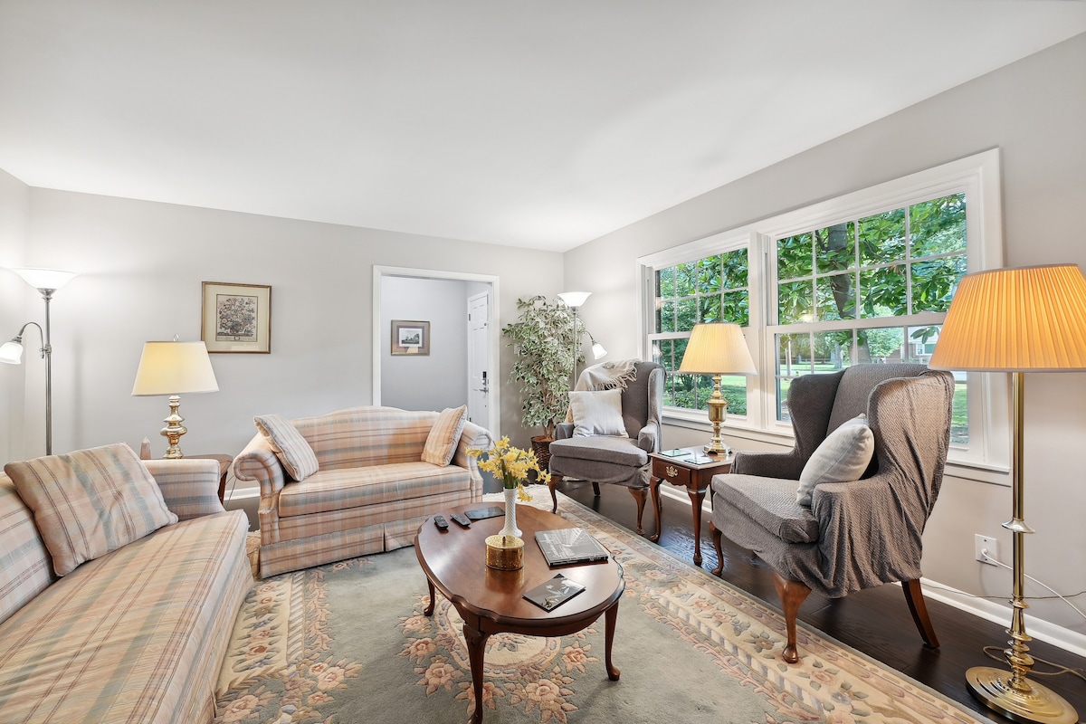 A bright living area features soft seating with a checked pattern, accompanied by a wooden coffee table. Large windows provide natural light and views of the outdoors. Two elegant armchairs are positioned next to a lamp, enhancing the comfortable atmosphere of the room.