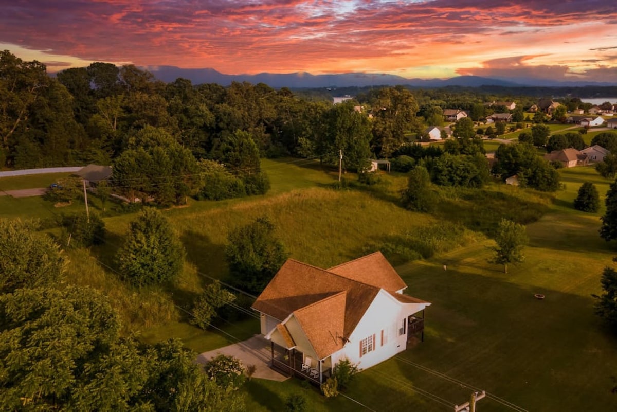 An aerial view showcases a charming home nestled in a spacious green landscape, surrounded by trees and rolling hills. A vibrant sunset illuminates the sky in shades of orange and pink, creating a serene backdrop for the peaceful countryside setting.