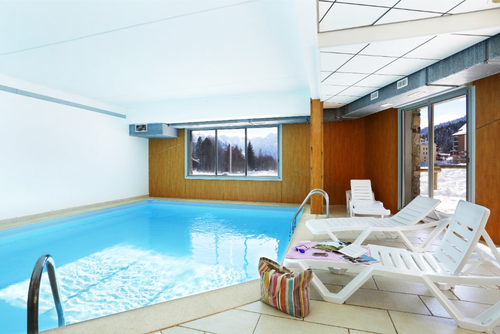 An indoor heated pool is featured, surrounded by large windows offering natural light and views of the outdoors. White lounge chairs are arranged around the pool area, providing seating for relaxation. The clean, minimalistic design creates a tranquil atmosphere.