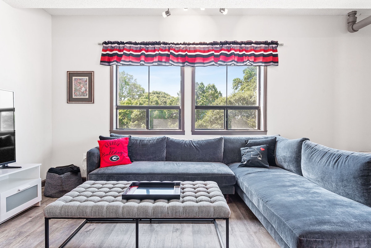 The living room features a spacious sectional sofa with soft upholstery, accented by a bulldog-themed pillow. Natural light enters through large windows, complemented by colorful drapes. A tufted coffee table is centrally placed, providing a functional surface for relaxation and enjoyment.