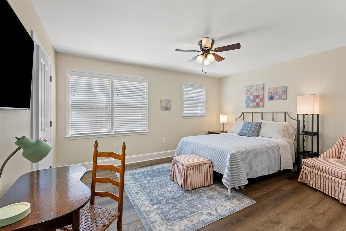 A bright bedroom showcases a spacious bed with a light-colored quilt, complemented by a decorative patterned area rug. Two windows provide natural light. A chair and desk are present, along with a small striped couch and a bedside lamp, creating a functional and inviting space.