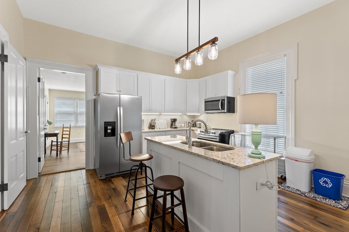 A modern kitchen features white cabinetry and granite countertops, complemented by stainless steel appliances. A central island with seating for two is illuminated by pendant lighting. Wood flooring extends throughout the space, creating a warm and inviting atmosphere.