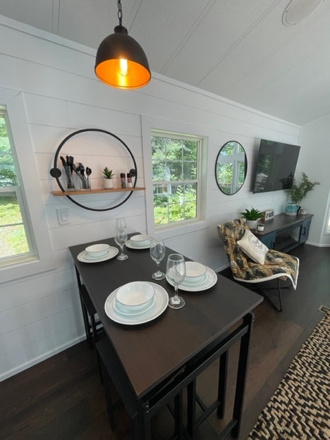 A modern dining area features a dark table set for four, complete with plates and glasses. Two round mirrors adorn the walls, while a shelf displays kitchen items. A cozy chair is positioned next to a TV, and light from a stylish pendant illuminates the space.