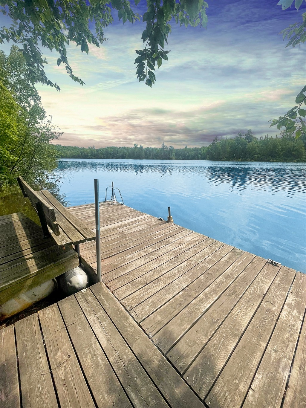 A wooden dock extends over calm water, surrounded by lush greenery and trees. Soft colors blend in the sky, reflecting peaceful sunset hues. A ladder leads into the water, providing access for swimming or relaxation by the lake.