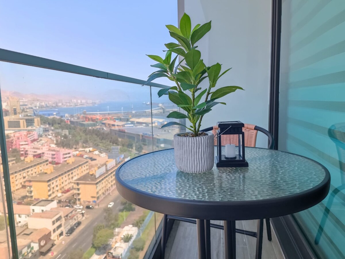 A balcony is furnished with a round table and two chairs, providing a view of the city and ocean. A potted plant and a lantern are placed on the table, complementing the serene outdoor space. The panoramic vista captures the landscape beneath.