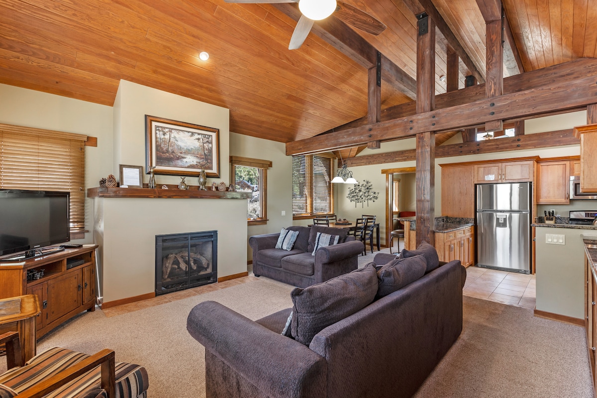 A spacious living area features a high wooden ceiling supported by exposed beams. A flat-screen TV is mounted above a stone fireplace, while a comfortable seating arrangement includes two dark couches. The room is designed for relaxation and entertaining, with open access to the dining area and kitchen.
