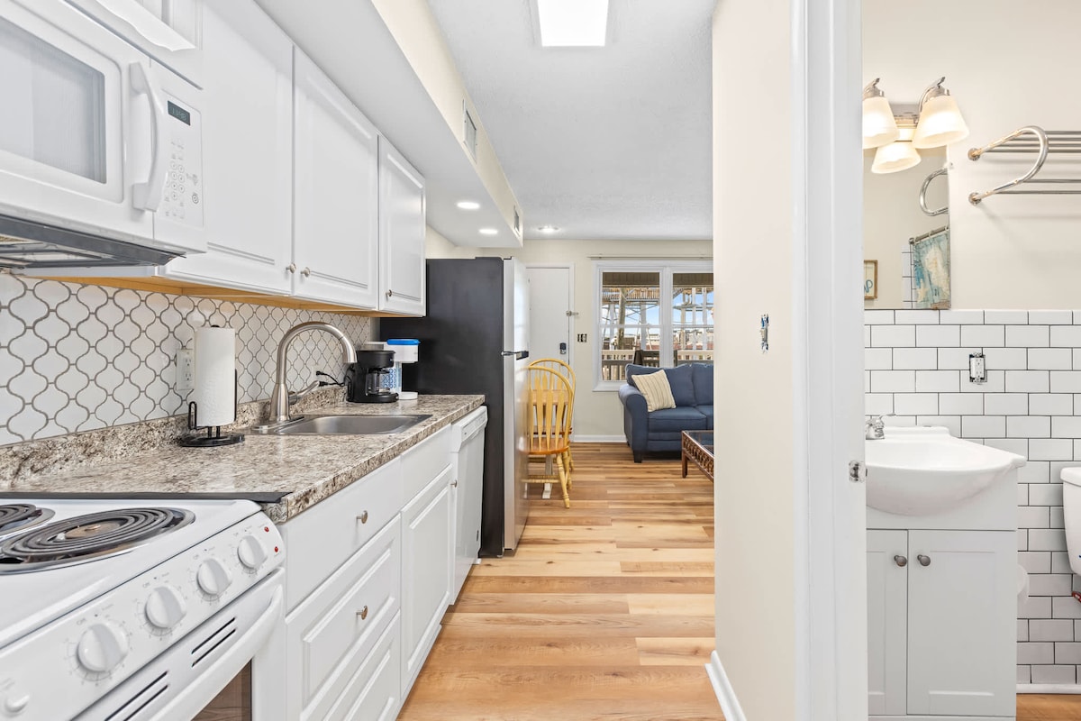 A bright kitchen area features white cabinetry and granite countertops, with essential appliances visible, including a microwave and stove. The space opens up to a cozy living area with a blue sofa and natural light streaming in from windows, creating an inviting flow.