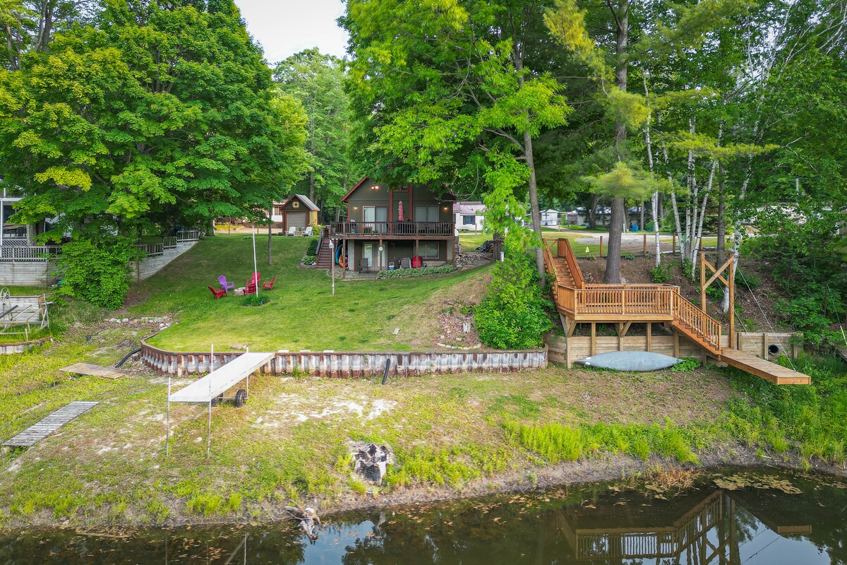 The cabin is set amid lush greenery, featuring a large deck that extends towards the water. A landscaped area includes a small dock for access to the Tittabawassee River. Red chairs are situated near the water's edge, inviting relaxation and enjoyment of the natural surroundings.