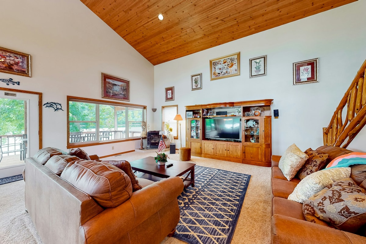 A spacious living area features a warm wooden ceiling and large windows overlooking the outdoors. Comfortable sofas with tasteful cushions are arranged around a central coffee table. A wooden entertainment unit houses a flat-screen television and decorative accents, creating a welcoming atmosphere.