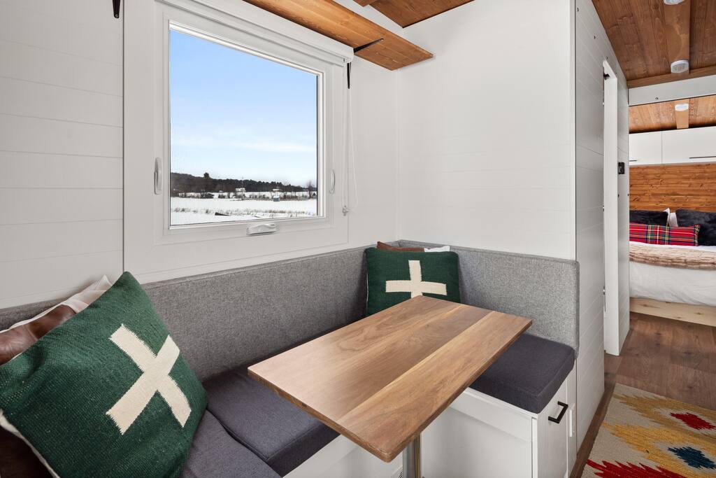 A cozy dining nook features a wooden table surrounded by upholstered benches. Two decorative pillows with a cross design add color to the neutral, light-colored space. Natural light fills the area through a window, offering views of the surrounding landscape.