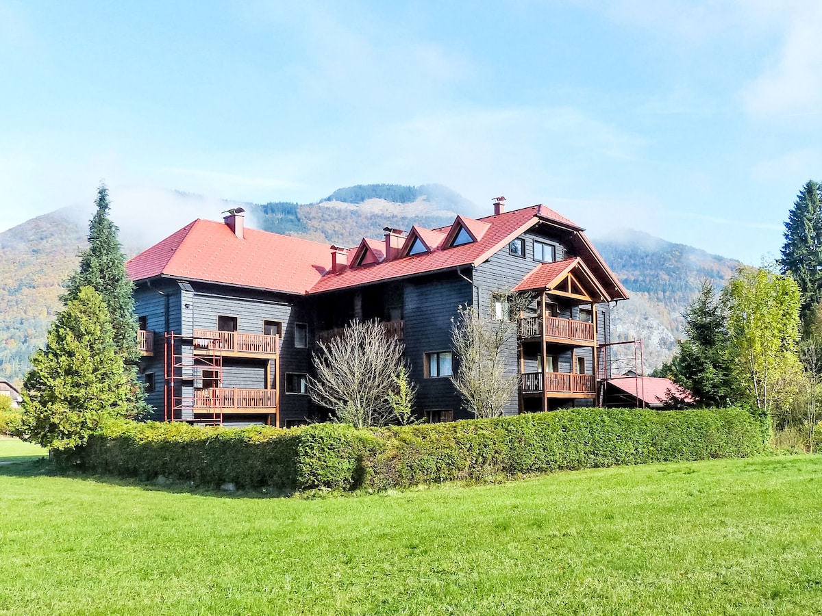 The three-story apartment building is set amidst lush greenery, featuring a dark wooden exterior with multiple balconies. A bright red roof complements the structure, while the surrounding landscape includes well-maintained hedges and open grassy areas, with mountains rising in the background.